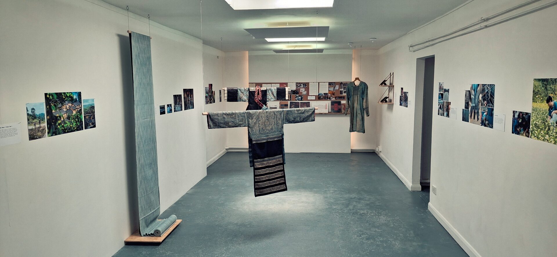 Installation view of a textile exhibition — hanging garments, indigo cloth and pinned research on the walls.