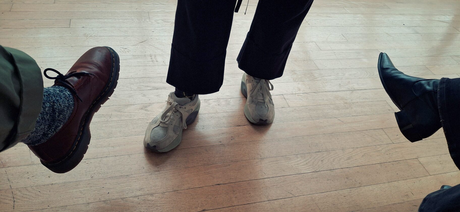 Four pairs of feet standing in a loose circle on a pale wooden floor — brown boots, sneakers, black boots.