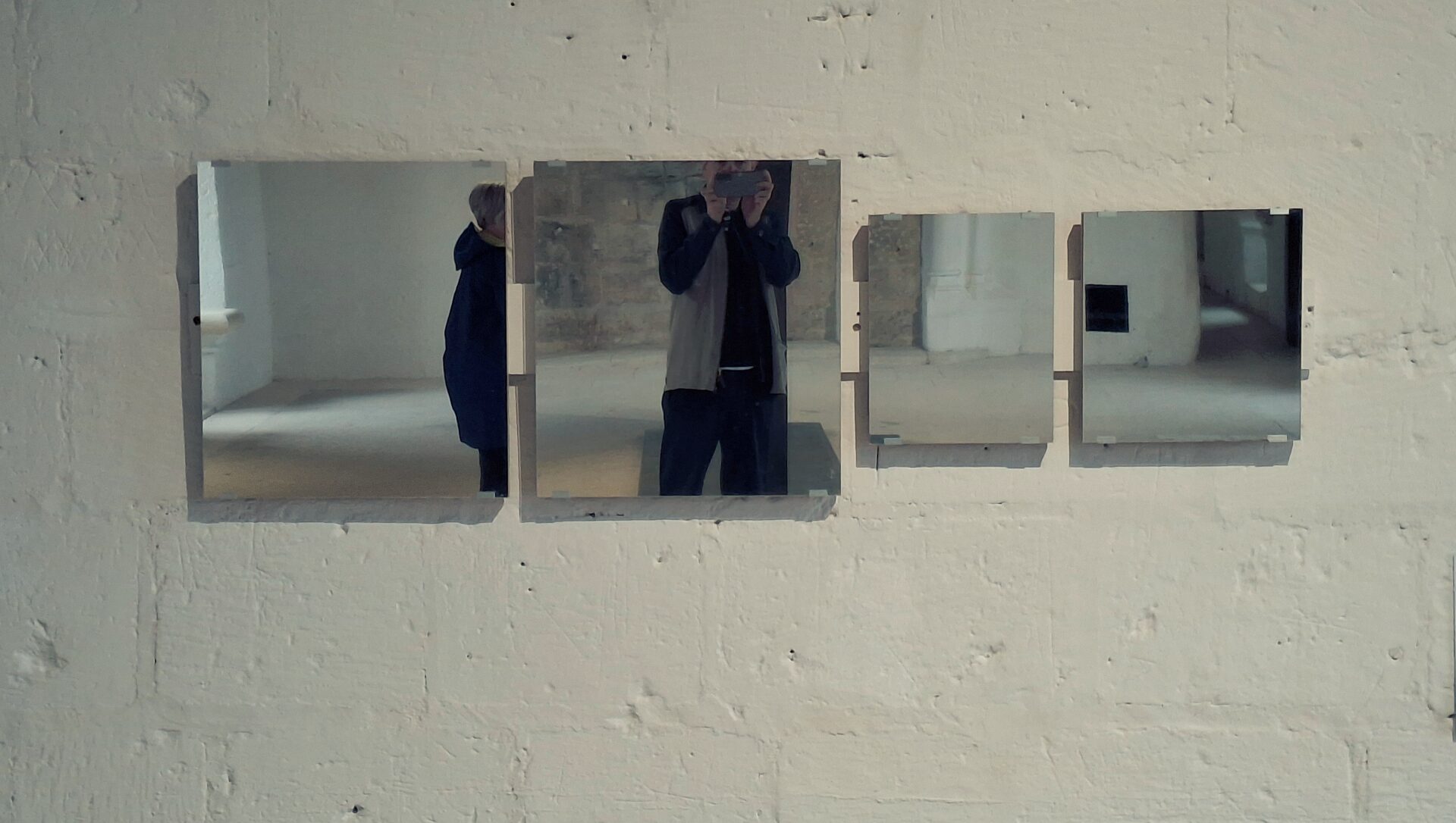 Four small square mirrors mounted on a whitewashed brick wall, each reflecting a different fragment of a room.