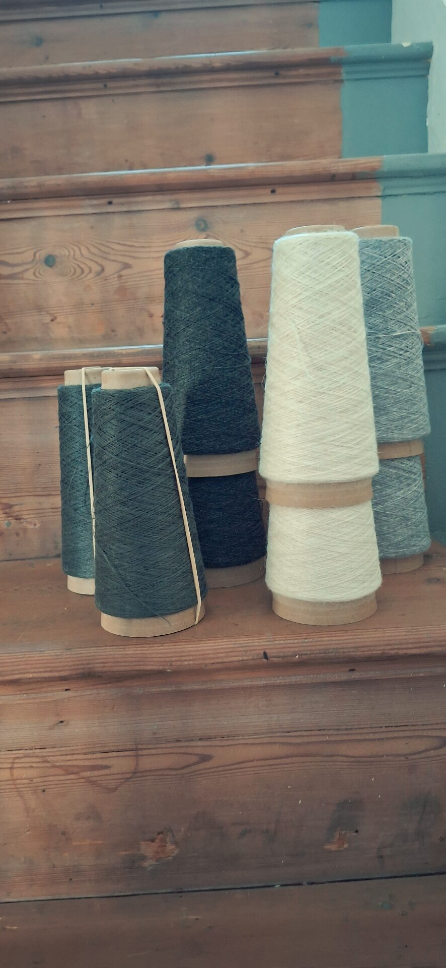 Cones of wool yarn in dark green, cream and grey, arranged on wooden stairs.
